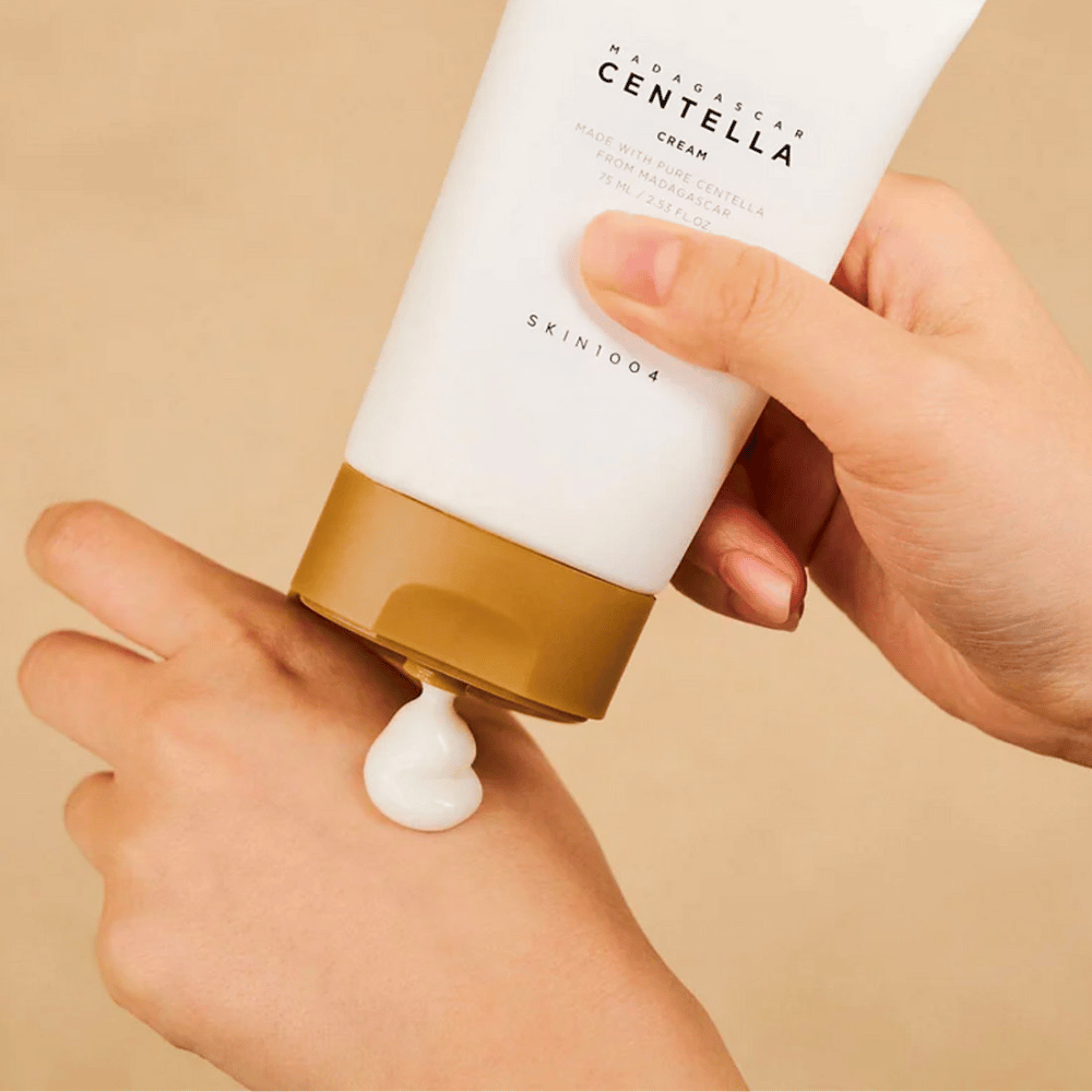Billedet viser en person, der holder en tube af "Madagascar Centella Cream" fra SKIN1004. Det er en hudcreme, der er fremstillet af ren Centella fra Madagaskar, i størrelsen 75 ml / 2.53 fl.oz. Cremen påføres på hånden og er beregnet til at give næring og fugt til huden.