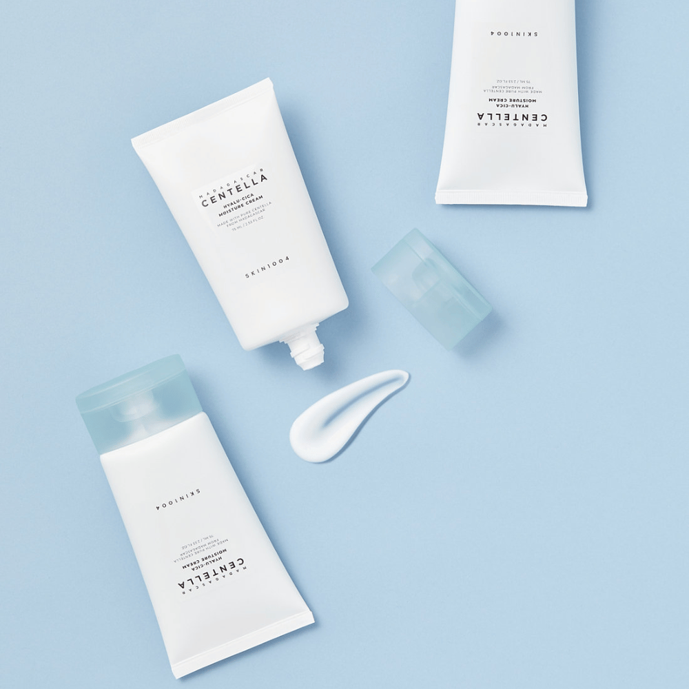 To hvide tuber med SKIN1004 Centella hudplejeprodukter, en klat af produkt og en klar blå kapsel, på en lys blå baggrund.