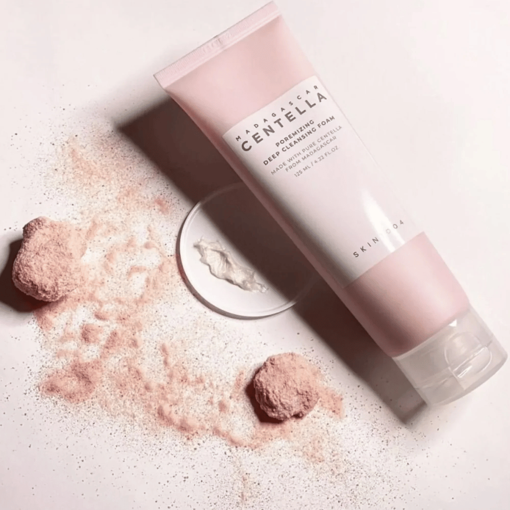 Madagascar Centella Poremizing Deep Cleansing Foam præsenteres i en lyserød tube. Tuben ligger på et lyst underlag sammen med lyserødt pulver, der giver en følelse af renhed og friskhed. Produktet er designet til at dybderense huden ved hjælp af ren centella fra Madagaskar.