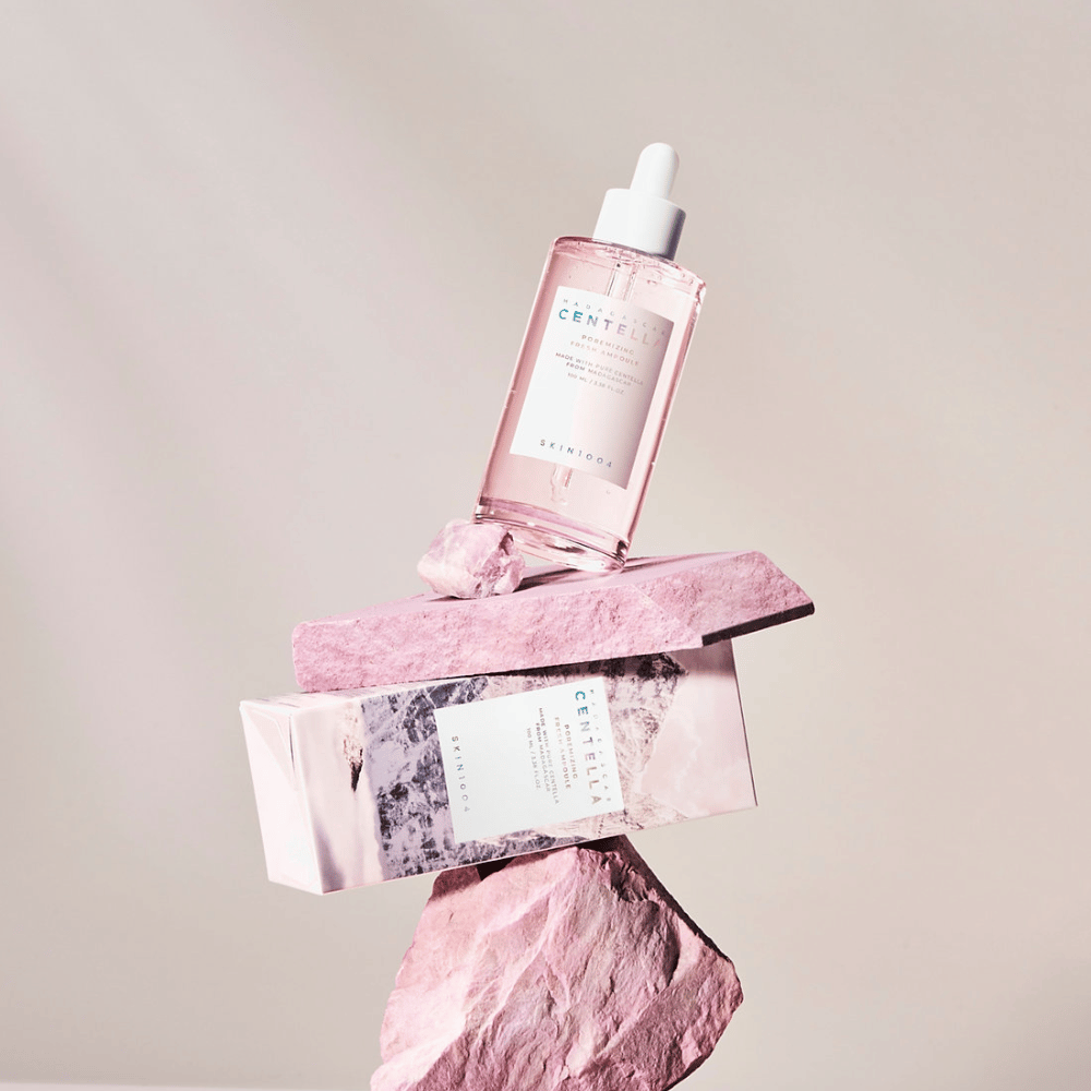 Gennemsigtig flaske med rosa Centella-ampul fra SKIN1004, balanceret på stenblok, med en papæske under, mod en neutral baggrund.