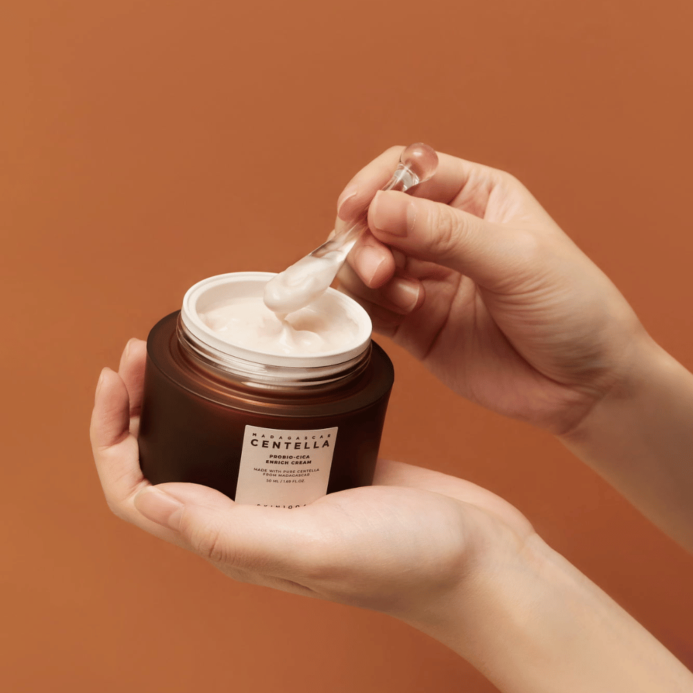 Hænder der holder en åben krukke af Madagascar Centella Probio-Cica Enrich Cream og en spatel med creme mod en ensartet terracottafarvet baggrund.