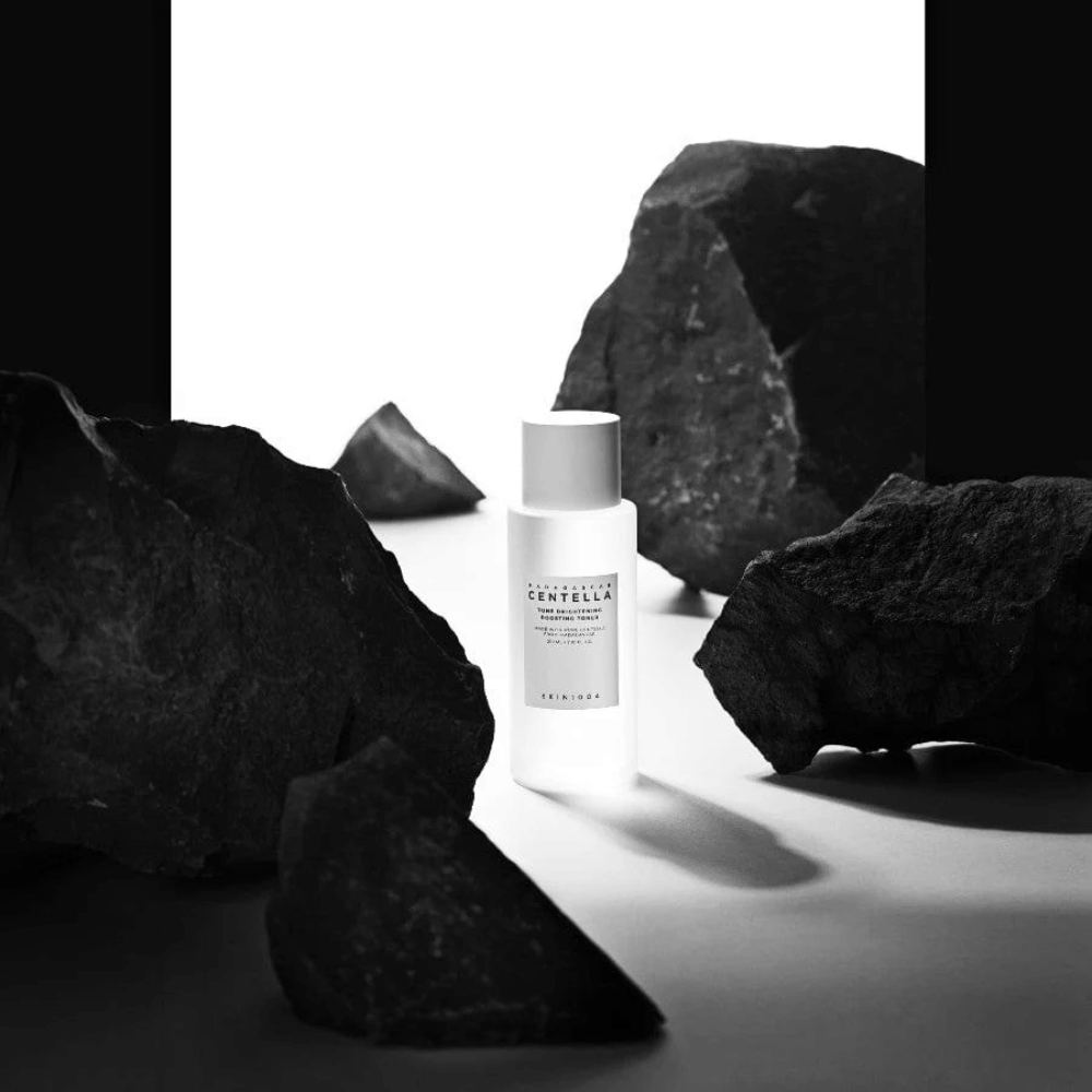 Hvid flaske SKIN1004 Madagascar Centella Tone Brightening Boosting Toner placeret dramatisk blandt store sorte sten mod hvid baggrund.