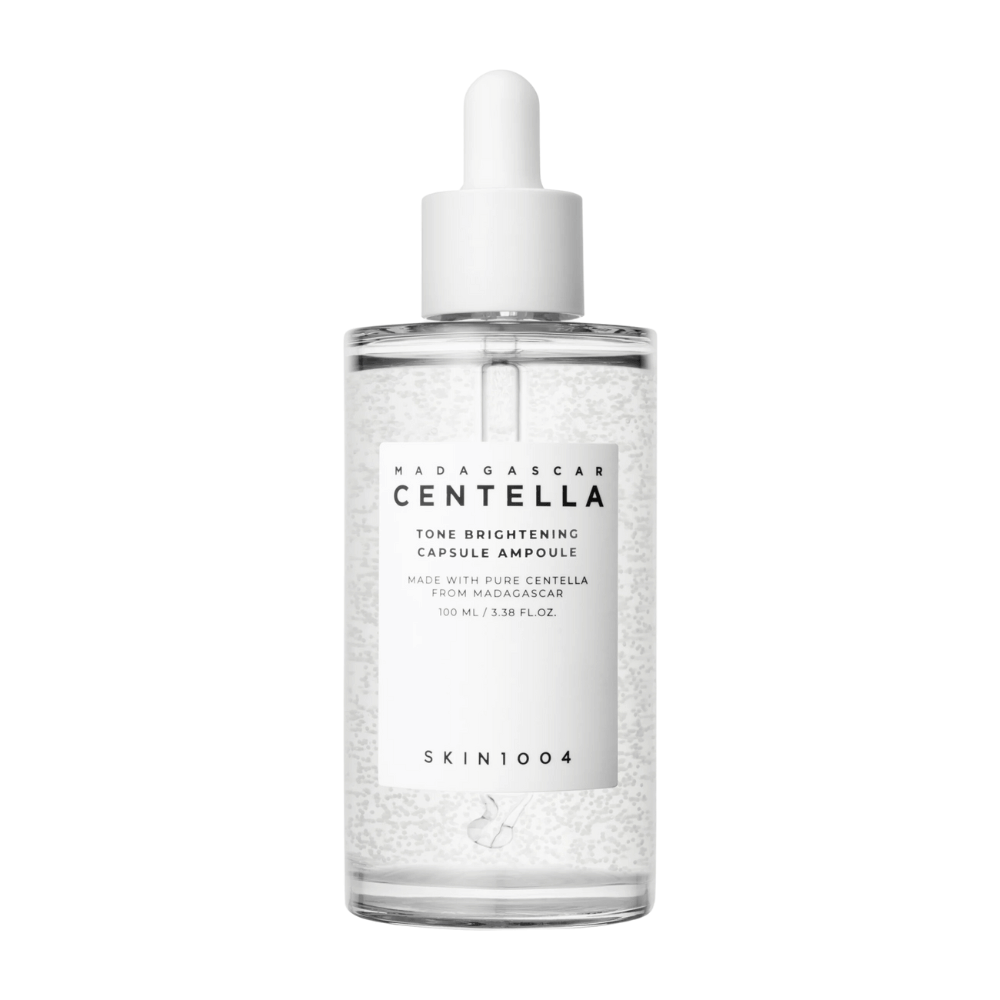Billedet viser en flaske af "SKIN1004 Madagascar Centella Tone Brightening Capsule Ampoule". Det er en gennemsigtig flaske med en hvid dråbeapplikator. På etiketten står der, at produktet indeholder rene centella-ingredienser fra Madagaskar. Flaskens indhold ser ud til at have små kapsler eller partikler i sig, hvilket kan være en del af dens lysnende hudplejeegenskaber. Flasken indeholder 100 ml / 3.38 fl.oz. af produktet.