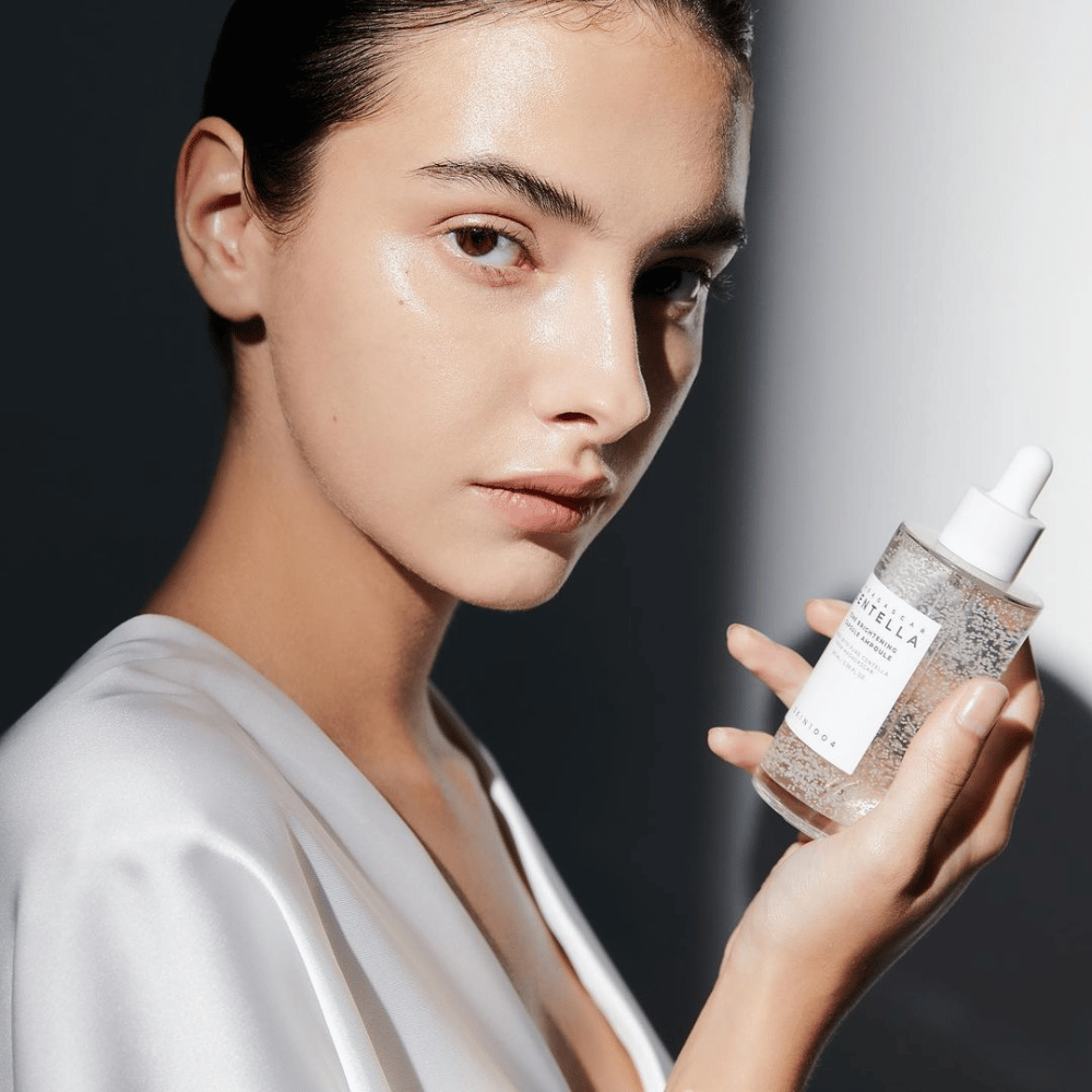På billedet ses en person, der holder en flaske med dråbeapplikator. Flasken er mærket "SKIN1004 Madagascar Centella Tone Brightening Capsule Ampoule". Personen har en skinnende hudtone, der reflekterer lyset, hvilket antyder en mulig effekt af produktet.