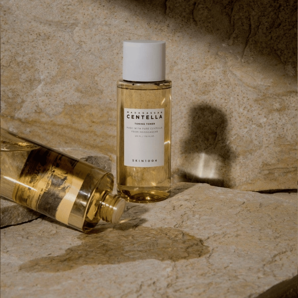 Billedet viser en flaske "Madagascar Centella Toning Toner" fra SKIN1004. Det er en transparent flaske fyldt med en gullig væske, sandsynligvis en toner til hudpleje. Det står, at den er lavet med ren centella fra Madagaskar, og indholdet er 210 ml / 7.10 fl.oz. Flasken og en liggende flaske er placeret på en stenbelagt overflade, hvilket giver en naturlig og minimalistisk følelse.