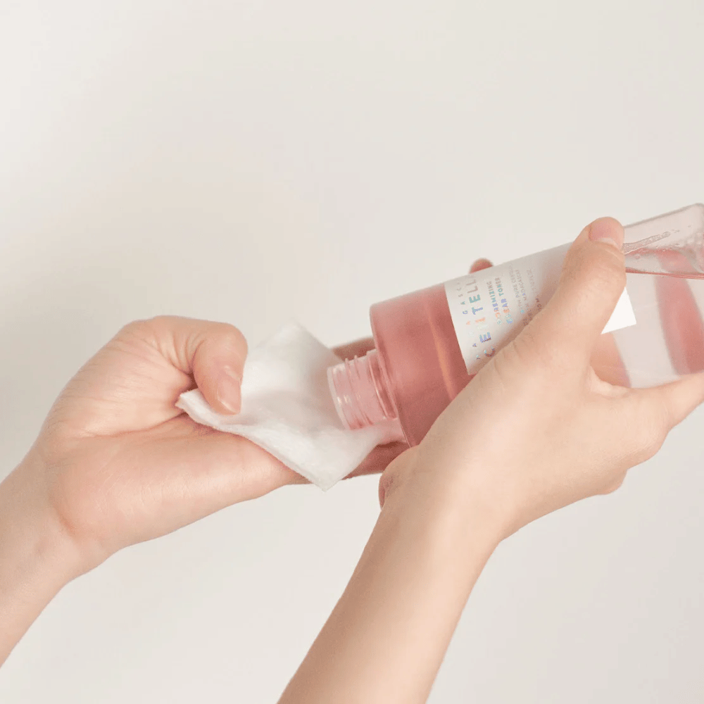 En person hælder Madagascar Centella Poremizing Clear Toner fra en transparent flaske på en vatrondel. Produktet, som er i en lyserød tone, bruges til at friske op og rense huden. Hænderne er synlige, hvilket viser påføringen af toneren.