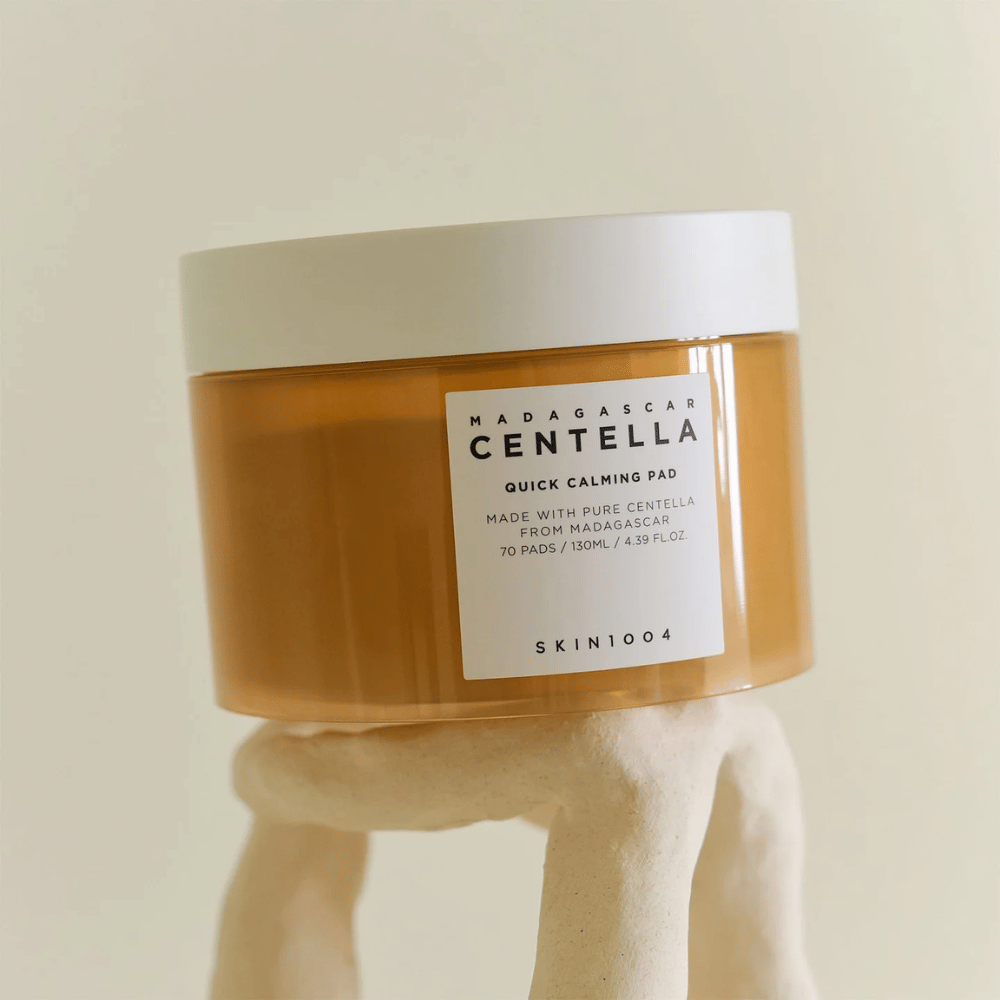 Bøtte med SKIN1004 Madagascar Centella Quick Calming Pad placeret på en skulpturel hvid piedestal – ravfarvet beholder med hvid etiket og låg.