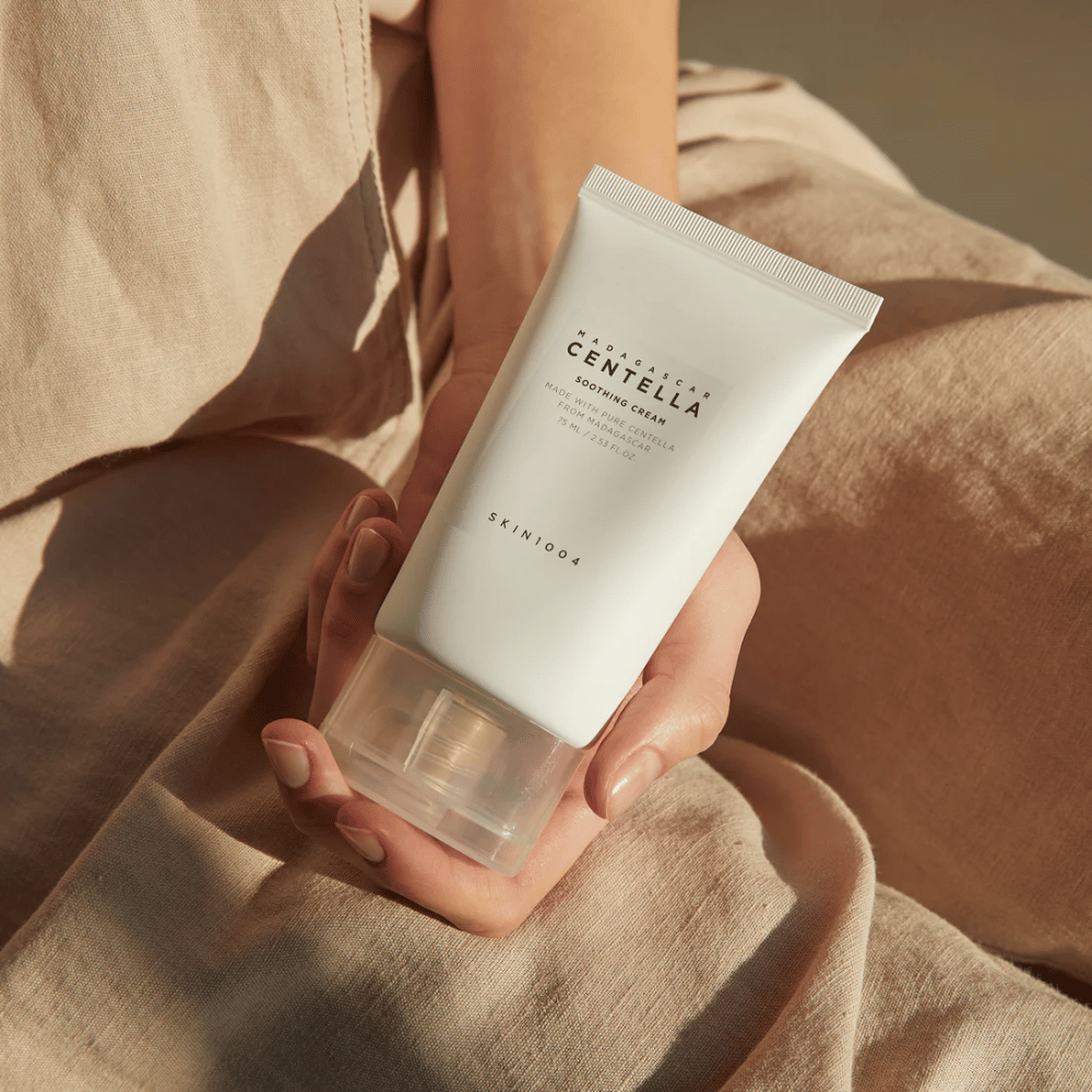 En person holder et hvidt rør af Madagascar Centella Soothing Cream fra SKIN1004. Røret har en gennemsigtig kork og tekst, der beskriver produktet som beroligende og fremstillet med ren Centella. Personen er klædt i lyse tøj.