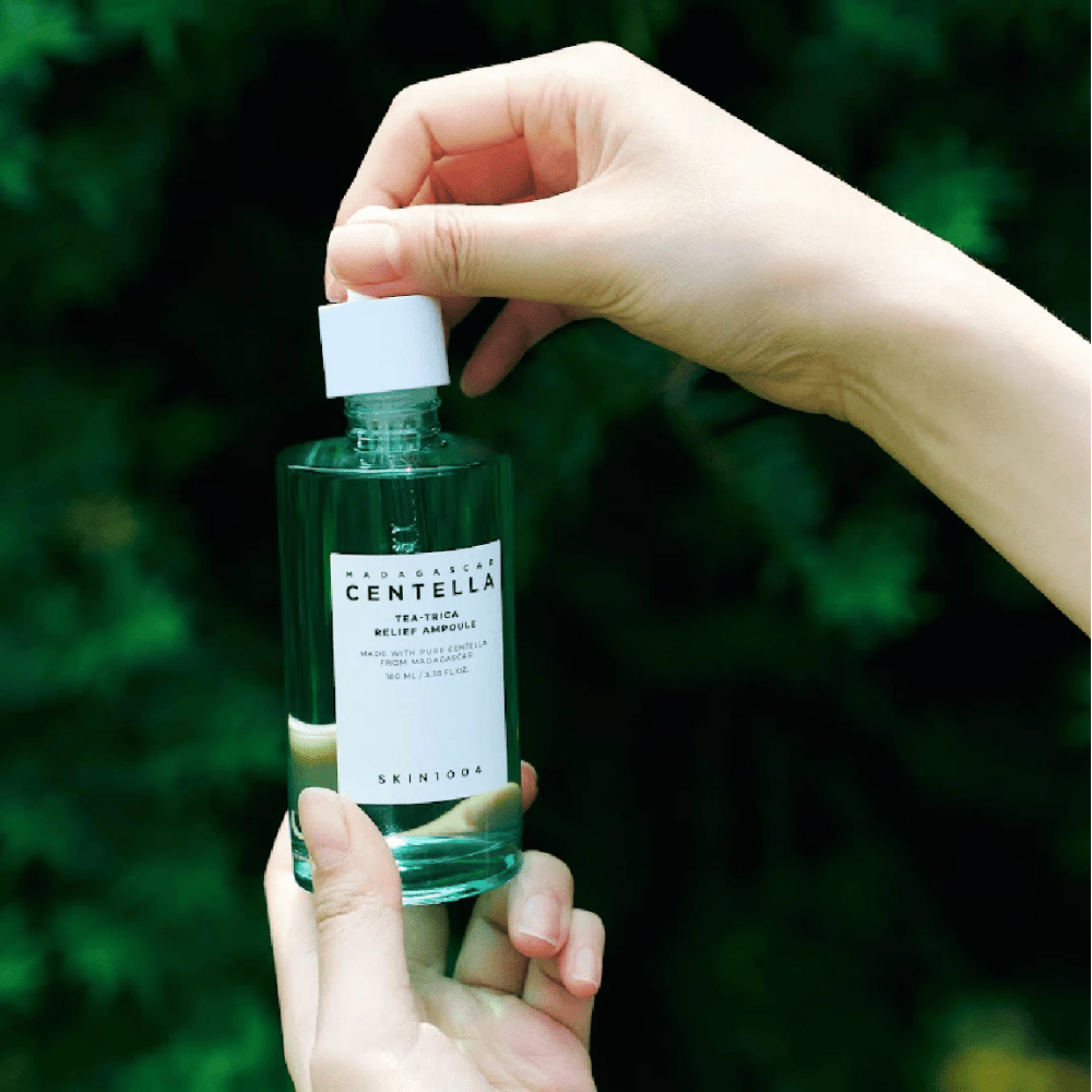 Person åbner SKIN1004 Madagascar Centella Tea-Trica Relief Ampoule – grøn glasflaske med hvid etiket holdes i hænder foran frodig baggrund.