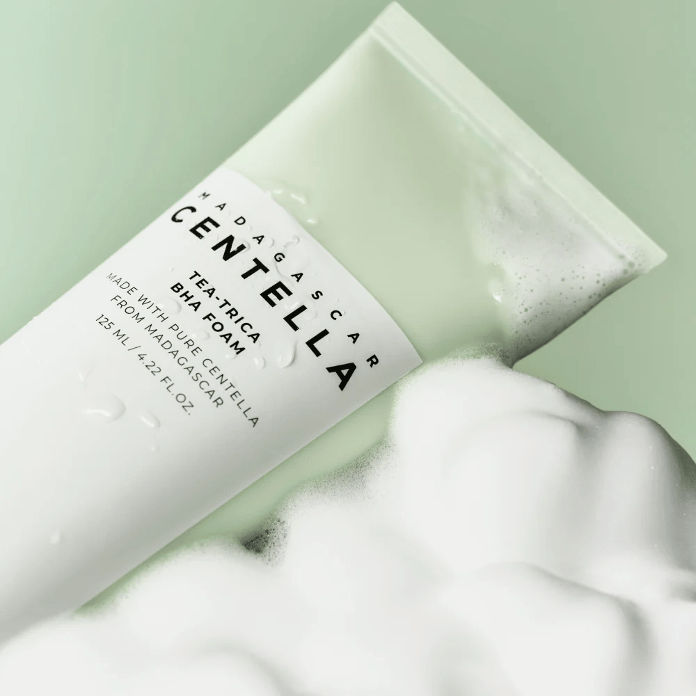 Et produkt af typen renseskum, "Madagascar Centella Tea-trica BHA Foam," er delvist synligt med skum rundt omkring. Produktet har et grønt og hvidt design, og teksten "Madagascar Centella" og "Tea-trica BHA Foam" er tydeligt synlige. Volumen er 125 ml.