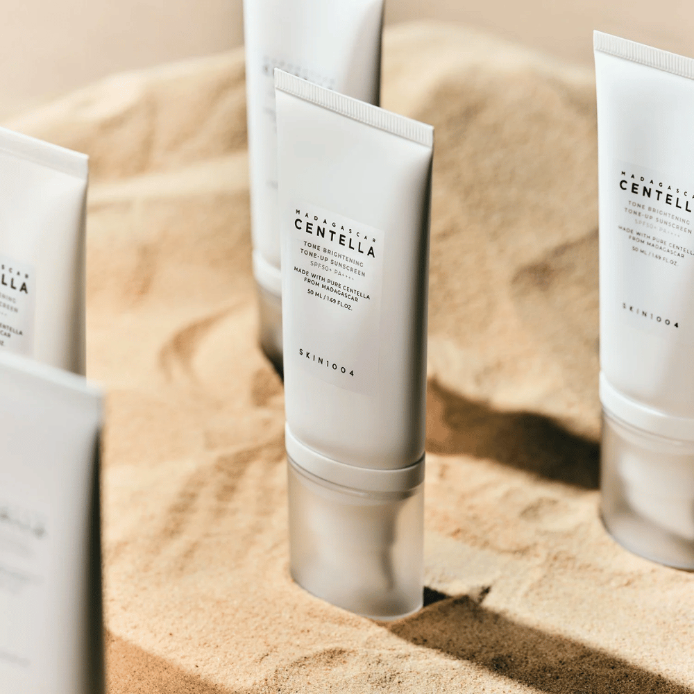 Flere tuber med Madagascar Centella Tone Brightening Tone-Up Sunscreen står i sandet. Tuben er hvid med sort tekst og indeholder 50 ml. Produktet har SPF 50+ PA++++ og er fremstillet med ren Centella fra Madagaskar.