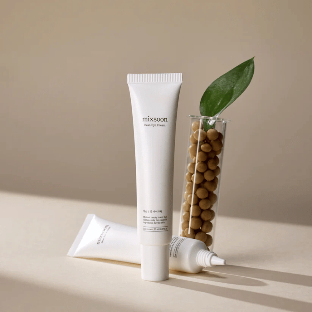To tuber Mixsoon Bean Eye Cream placeret ved siden af et reagensglas fyldt med sojabønner og et grønt planteblad, på en beige overflade med skyggespil.
