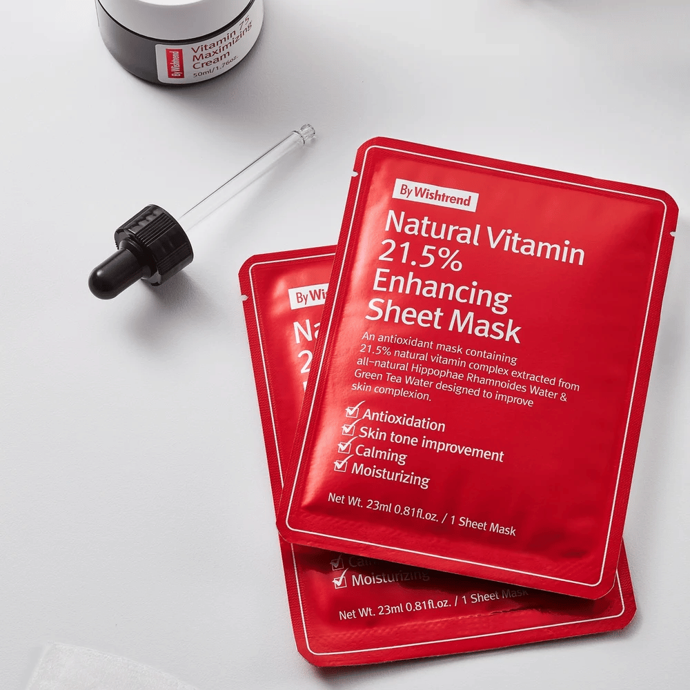 By Wishtrend Natural Vitamin 21.5% Enhancing Sheet mask i røde pakker, placeret på en hvid overflade sammen med en pipette og en krukke ansigtscreme.