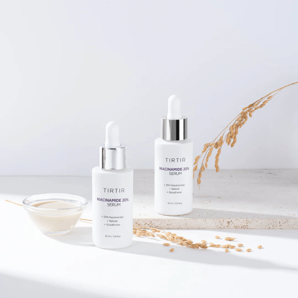 To flasker af Tirtirs "Niacinamide 20% Serum" vises på en lys overflade sammen med en lille skål med en cremet substans og nogle strå af tørrede planter. Serummet indeholder 20 % niacinamid, retinol og glutation, hvilket bidrager til at forbedre hudens udseende og tekstur.