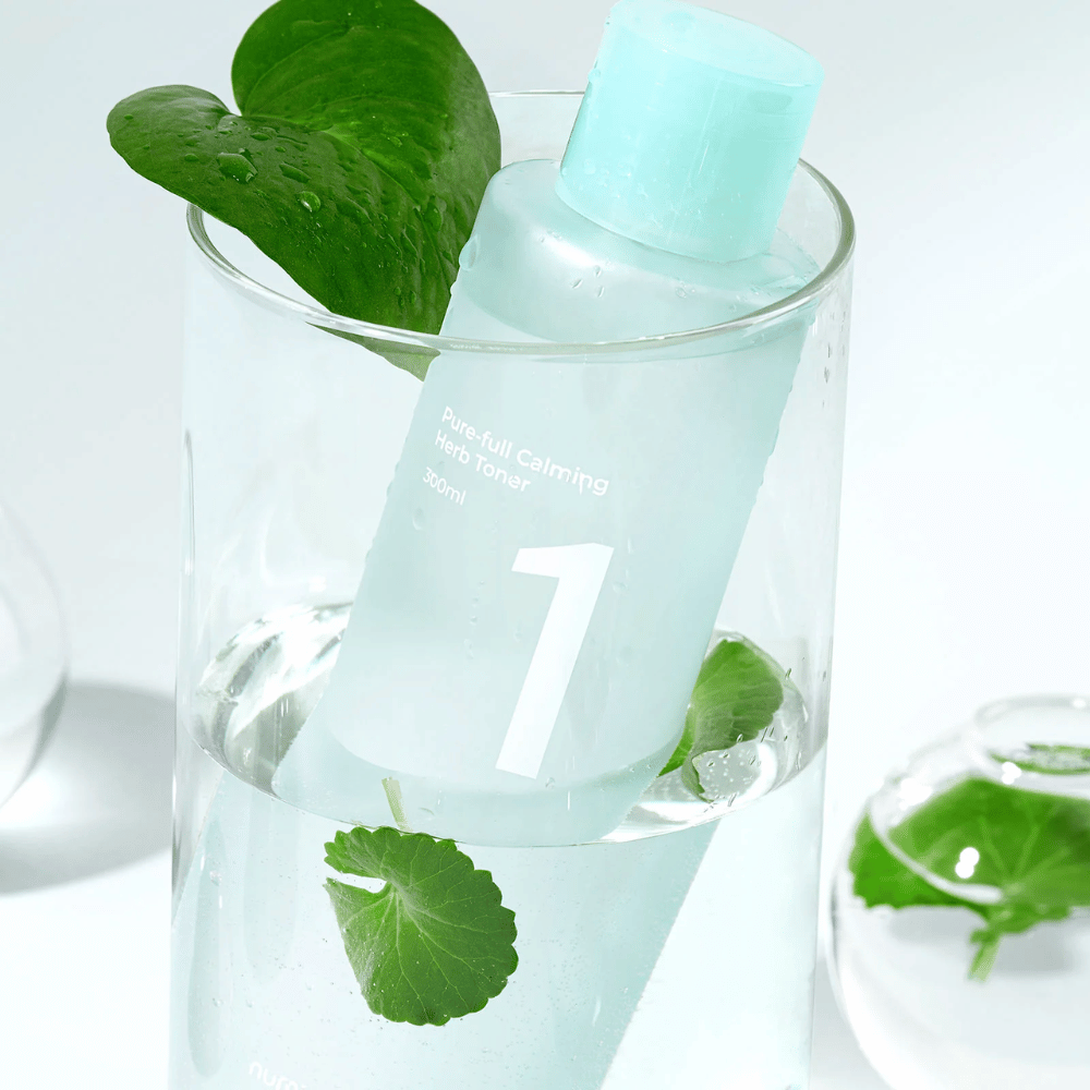 En flaske med No.1 Pure-Full Calming Herb Toner placeret i et glas med vand og grønne blade. Flasken har en lyseblå kork og en etiket med teksten "Pure-full Calming Herb Toner" samt "300ml". Produktet ser frisk ud med vand og vegetation i baggrunden, hvilket giver en beroligende og naturlig følelse.