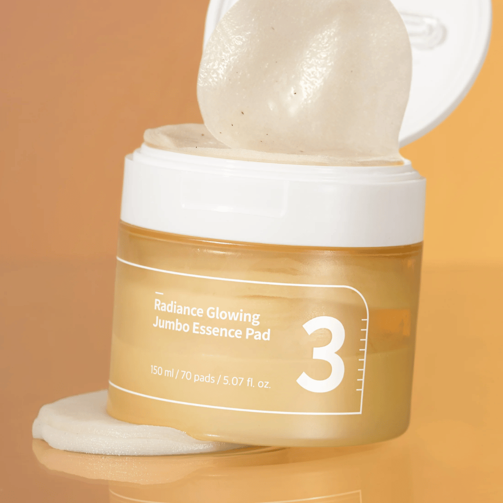 Åben krukke med numbuzin No. 3 Radiance Glowing Jumbo Essence Pad, en pad ses lænet mod kanten, baggrund i orange tone.