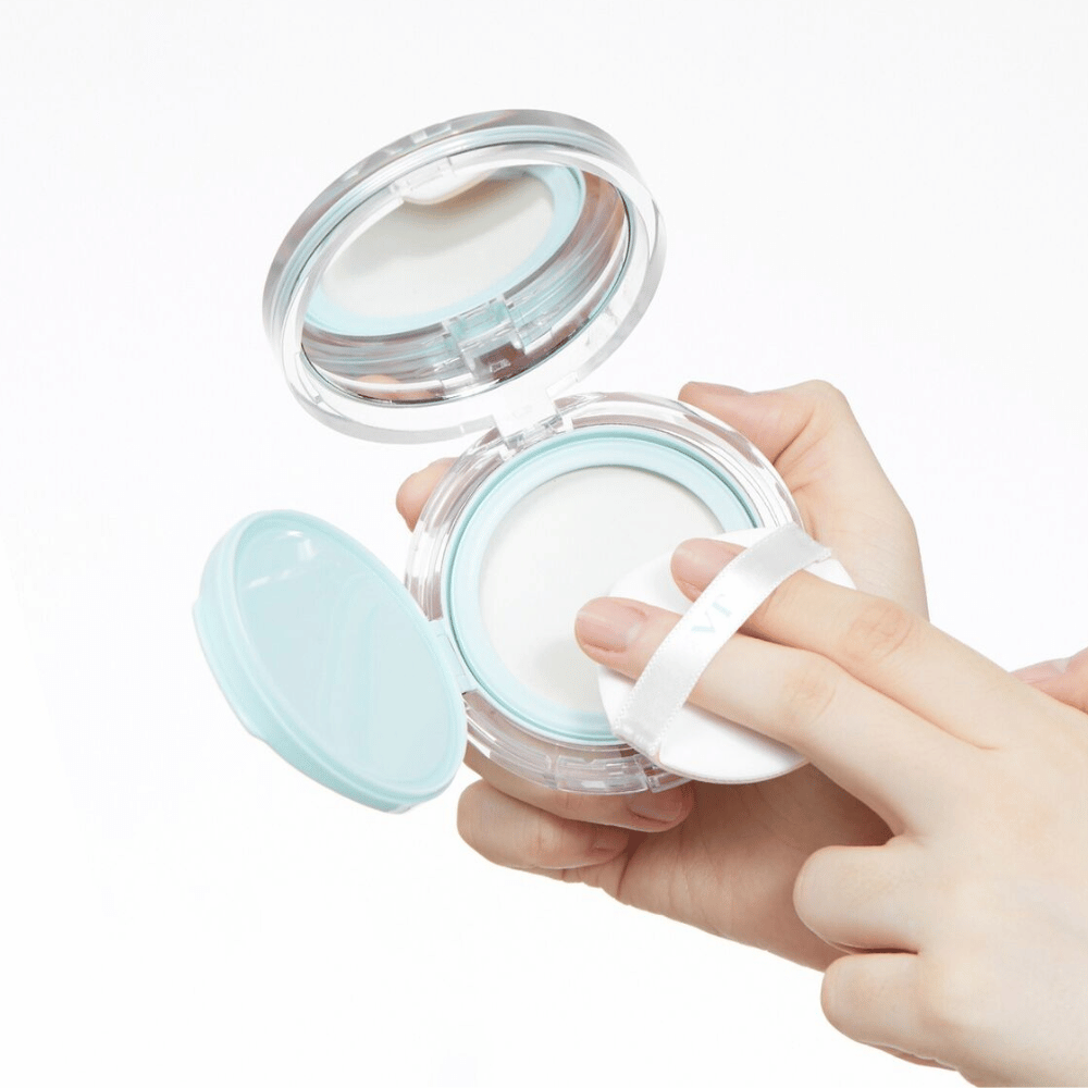En hånd holder en åben krukke med VT PDRN Essence Glow Sun Pact. Puffen er på vej til at påføre pudderet, som har en hvid overflade og en blå kant rundt om krukken.