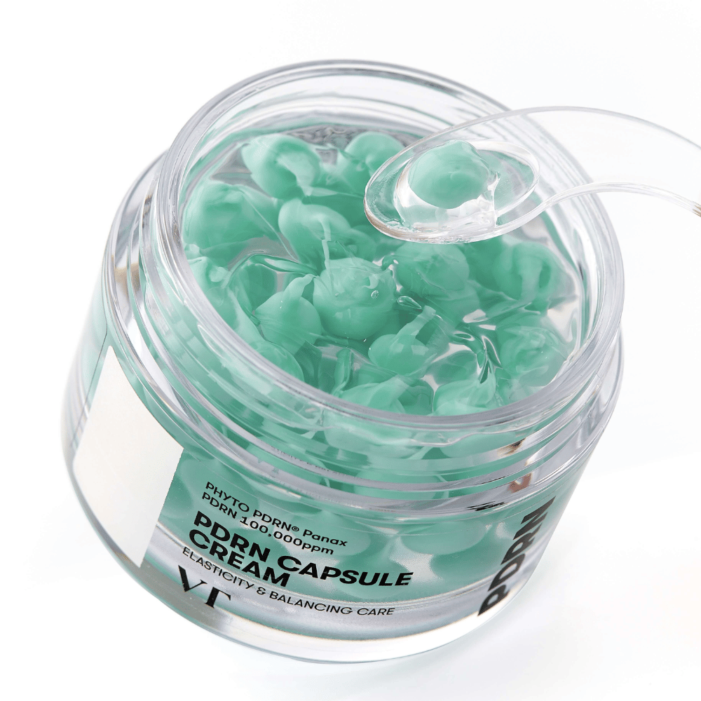 Nærbillede af en åben krukke med VT PDRN Capsule Cream. Indholdet består af små grønne kapsler i en gennemsigtig gel, med en medfølgende spatel til påføring. Teksten på krukken lyder "Elasticity & Balancing Care".