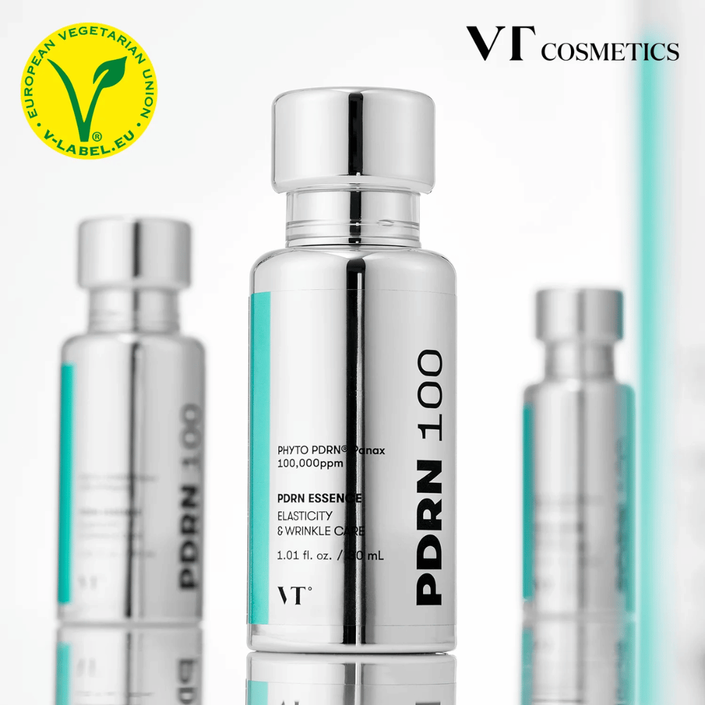 Flere sølvfarvede flasker af VT PDRN 100 Essence står på række, med tydelig mærkning om 100.000 ppm PDRN og fokus på elasticitet og rynkepleje. Øverst ses logoet for VT Cosmetics og et V-label for vegetabilsk indhold.