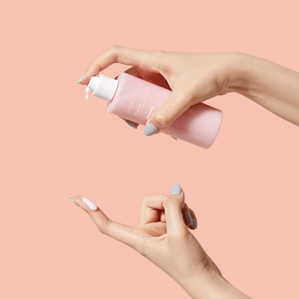 Billedet viser nogens hænder, der holder en flaske med ANUA Peach 77% Niacin Conditioning Milk. Dette hudplejeprodukt ser ud til at have en mælket konsistens og er designet til at pleje og forbedre hudens tilstand.