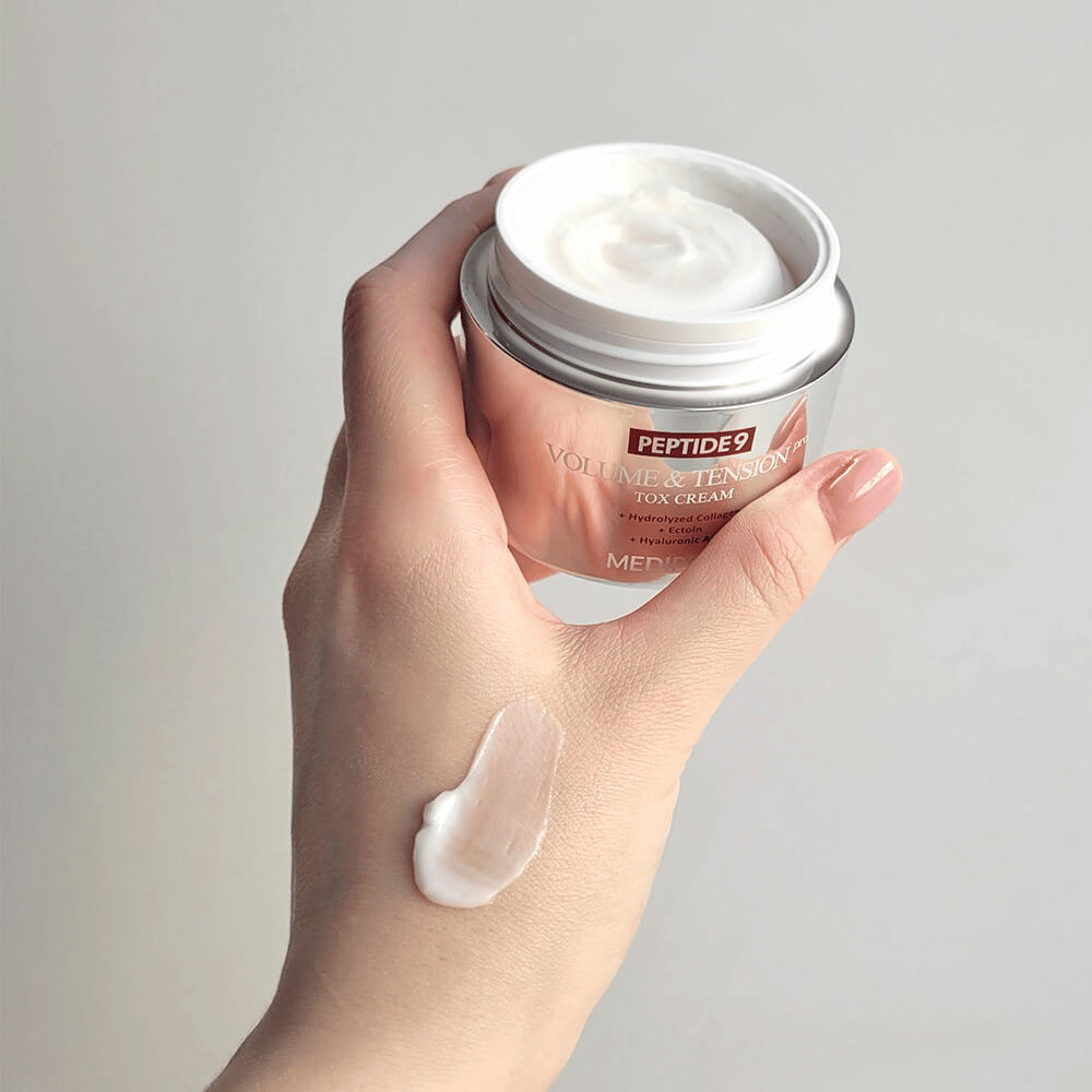 Hånd holder i MEDI-PEEL Peptide 9 Volume & Tension Tox Cream med en klat creme på håndryggen. Den silkebløde formel fugter og giver huden fasthed og spændstighed.