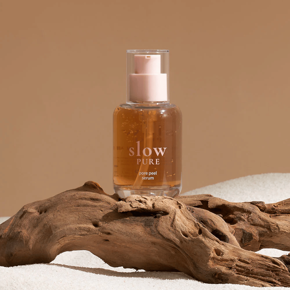 Slow Pure Pore Peel Serum placeret på sand med trædetalje i baggrunden, symbol for naturlig hudpleje.