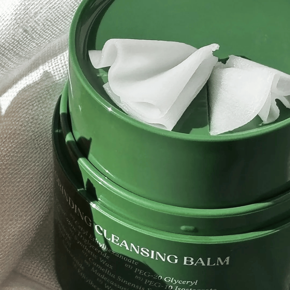 Billedet viser en grøn krukke med "Cleansing Balm", som er åbnet for at afsløre den medfølgende spatel og det bløde indhold. Den cremede konsistens reflekterer lyset, hvilket giver en følelse af luksus og effektivitet i rengøringen.