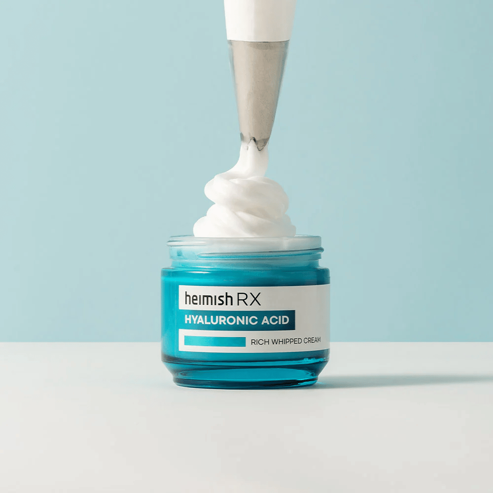 Heimish RX Hyaluronic Acid Rich Whipped Cream i en blå glasbeholder, hvor en sprøjtepose påfører den luftige creme. En fugtgivende ansigtscreme med hyaluronsyre for intensiv fugt og en blød, fyldig hud.