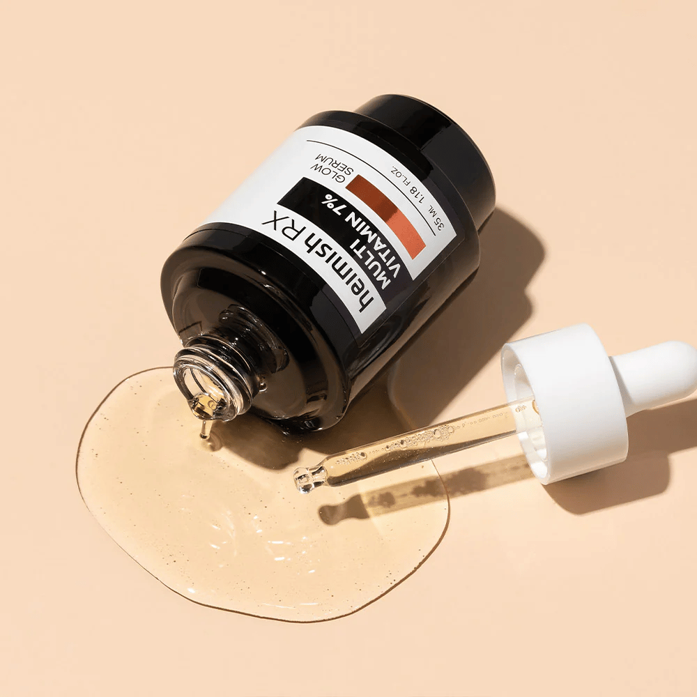 Heimish RX Multi Vitamin 7% Glow Serum liggende på en beige baggrund med serum spredt rundt om flasken og en pipette fyldt med produkt, hvilket fremhæver dens næringsrige formel.