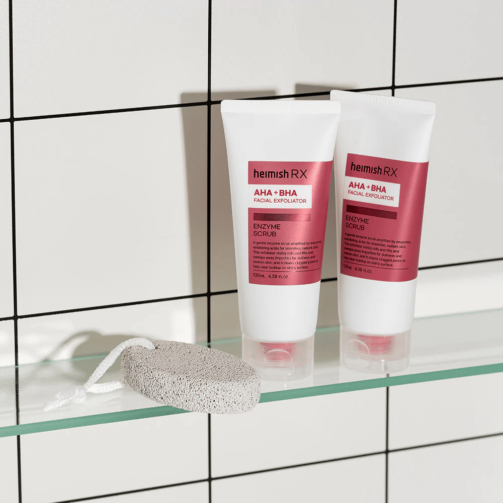To tuber Heimish RX AHA + BHA Facial Exfoliator placeret på en glashylde i et badeværelse med hvid flisevæg. Ved siden af produkterne ligger en pimpsten med en hvid snor. Et stilrent og minimalistisk hudplejebillede.
