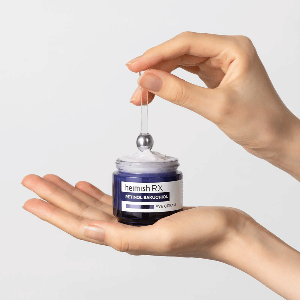 Heimish RX Retinol Bakuchiol Eye Cream i en elegant blå krukke, løftes op med en metalapplikator. En øjencreme med retinol og bakuchiol for at reducere fine linjer og give en fastere hud.