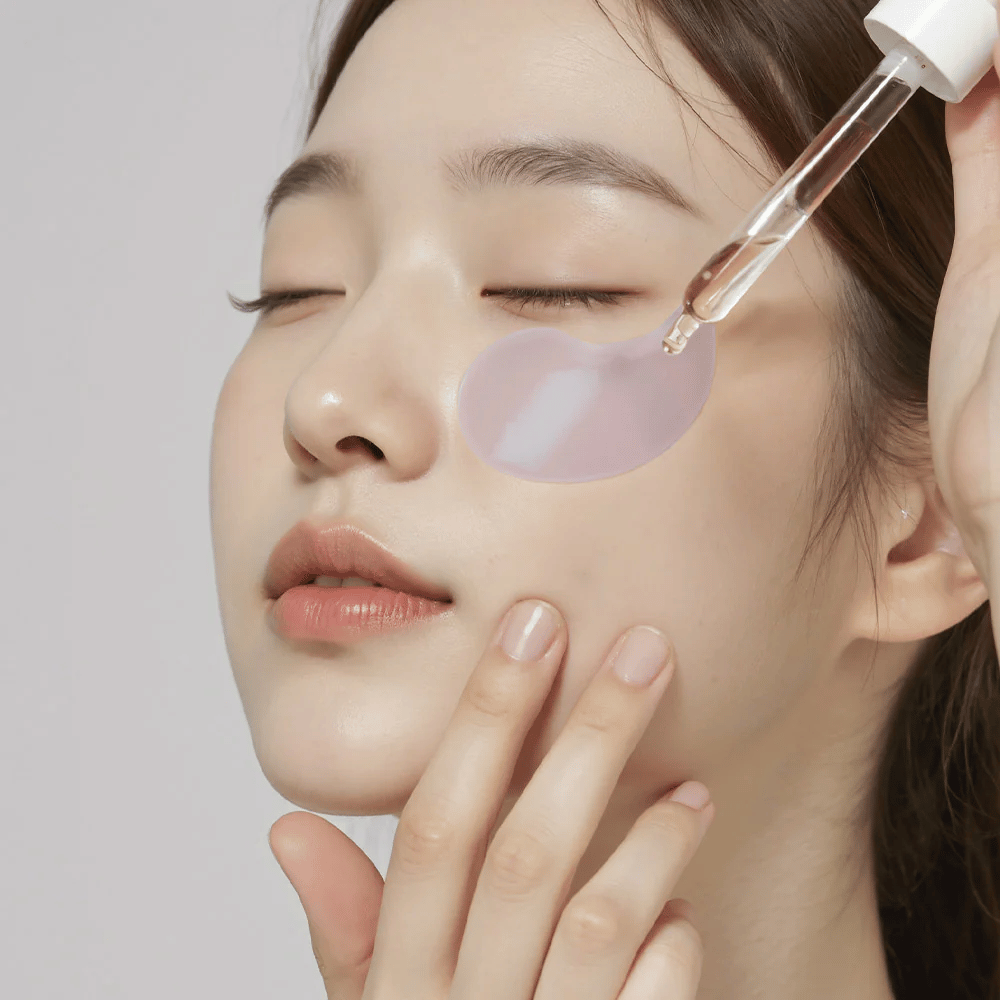 En kvinde med en lilla hydrogel-øjenlap påfører serum under øjet med en pipette. Produktet fugter og plejer den følsomme hud omkring øjnene.