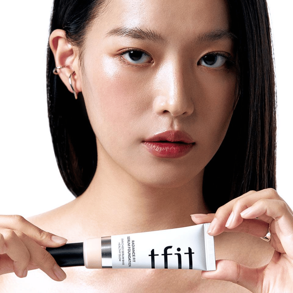 Model med glansfuld, jævn hud holder i TFIT Radiance Fit Serum Foundation, hvilket fremhæver produktets naturlige finish og glans.