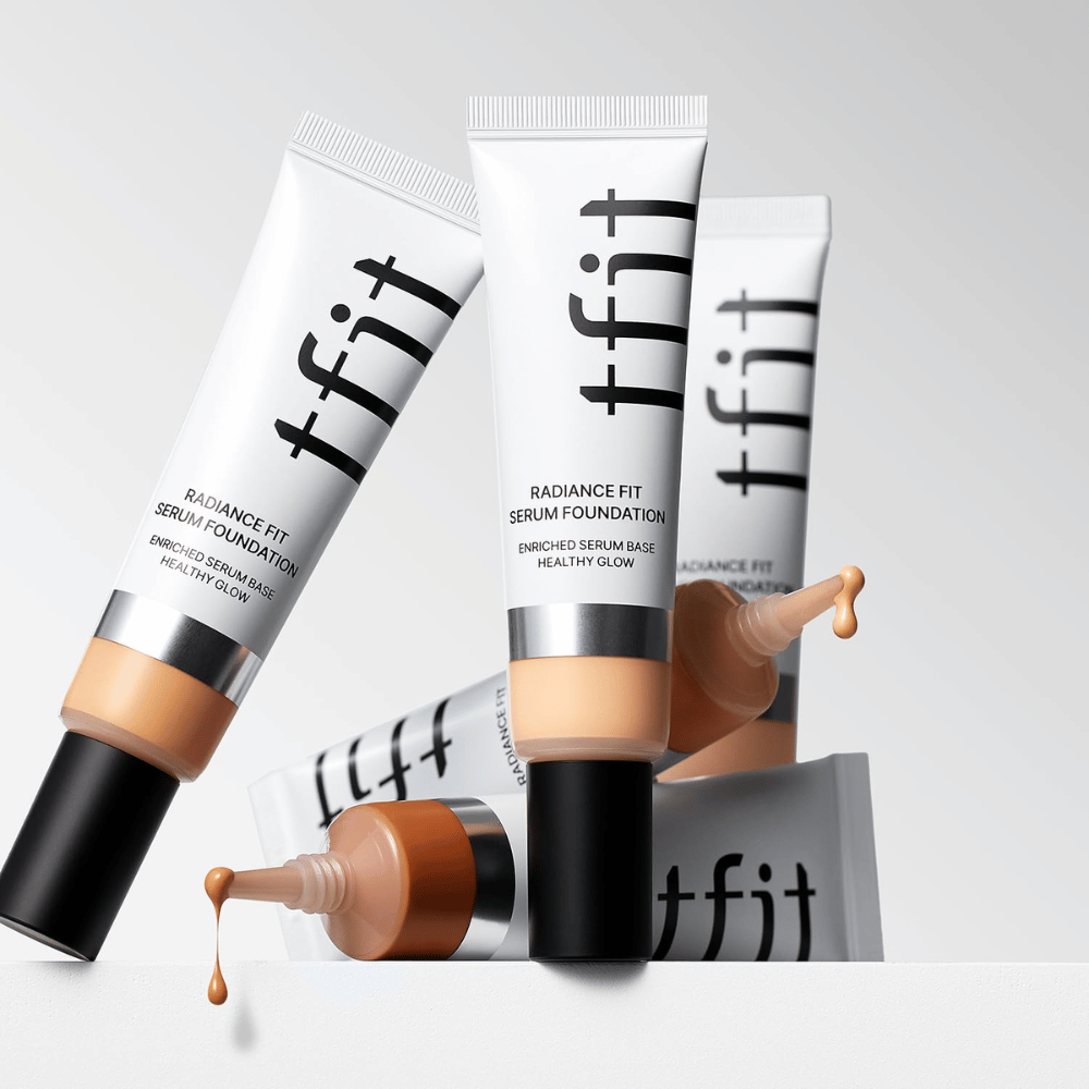 Flere tuber af TFIT Radiance Fit Serum Foundation i forskellige nuancer, hvoraf en med foundation som drypper ud, mod lysegrå baggrund.