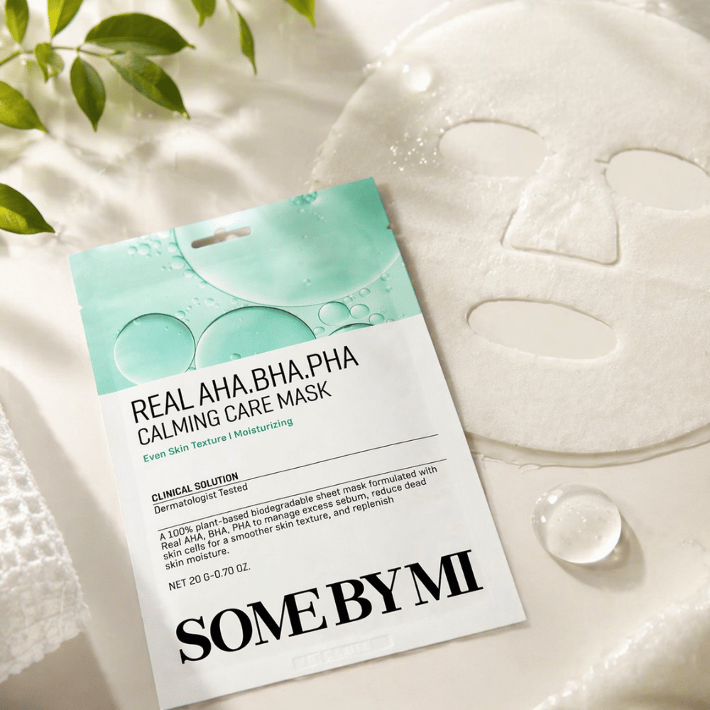 SOME BY MI Real AHA.BHA.PHA Calming Care Mask vises sammen med den udfoldede sheetmaske. Rundt om produktet ses fugtdråber og grønne blade, som forstærker følelsen af fugt, beroligende pleje og skånsom eksfoliering. Billedet fremhæver maskens fokus på en mere ensartet hudtekstur og forbedret fugtbalance.