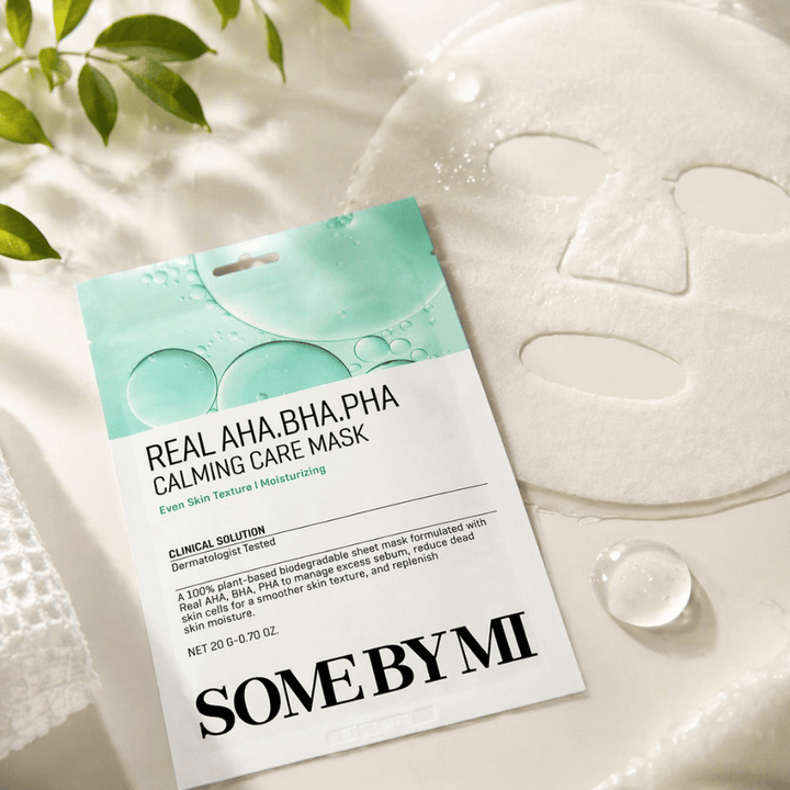 SOME BY MI Real AHA.BHA.PHA Calming Care Mask vises sammen med den udfoldede sheetmaske. Rundt om produktet ses fugtdråber og grønne blade, som forstærker følelsen af fugt, beroligende pleje og skånsom eksfoliering. Billedet fremhæver maskens fokus på en mere ensartet hudtekstur og forbedret fugtbalance.