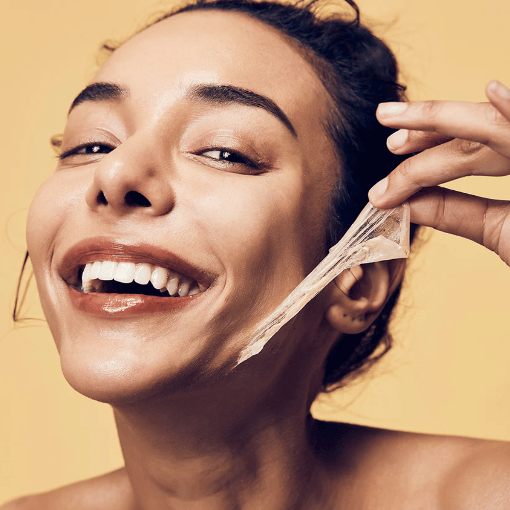 Billedet viser en kvinde med et stort smil, der er ved at fjerne en peel-off maske fra sit ansigt. Kvinden ser glad og tilfreds ud, mens hun bruger skønhedsproduktet, hvilket formidler en følelse af velvære og hudpleje.