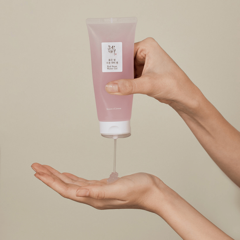 Billedet viser en person, der holder en tube af "Red Bean Water Gel" fra Beauty of Joseon og klemmer en gennemsigtig gel ud i sin hånd. Produktet ser ud til at være beregnet til hudpleje og har en attraktiv, blød lyserød emballage.