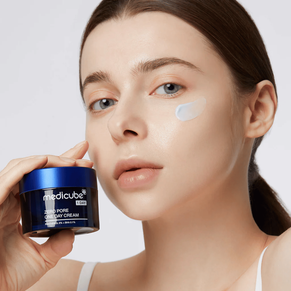 Kvinde med et naturligt look holder Medicube Zero Pore One Day Cream. En lille mængde creme er påført hendes kind. Produktet er designet til at forbedre hudens struktur og minimere porestørrelse, med en let og fugtgivende formel.