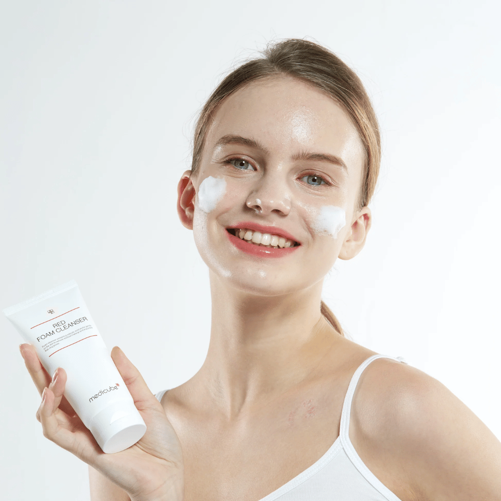 Ung kvinde med renseskum i ansigtet holder en tube af Medicube Red Foam Cleanser og smiler til kameraet. Hun bærer en hvid top, og baggrunden er lys og enkel.