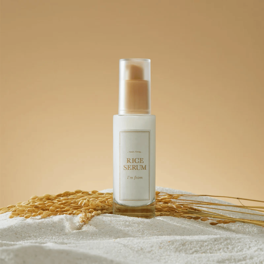Flaske med "I'm from" Rice Serum, omgivet af riskorn og risaks mod en varm baggrund.