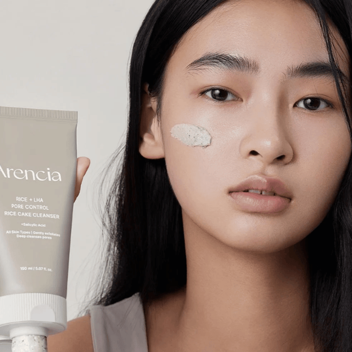 Model der holder Arencia Rice + LHA Pore Control Rice Cake Cleanser med påført produkt på kinden, illustrerer mild men effektiv porerens til daglig brug.