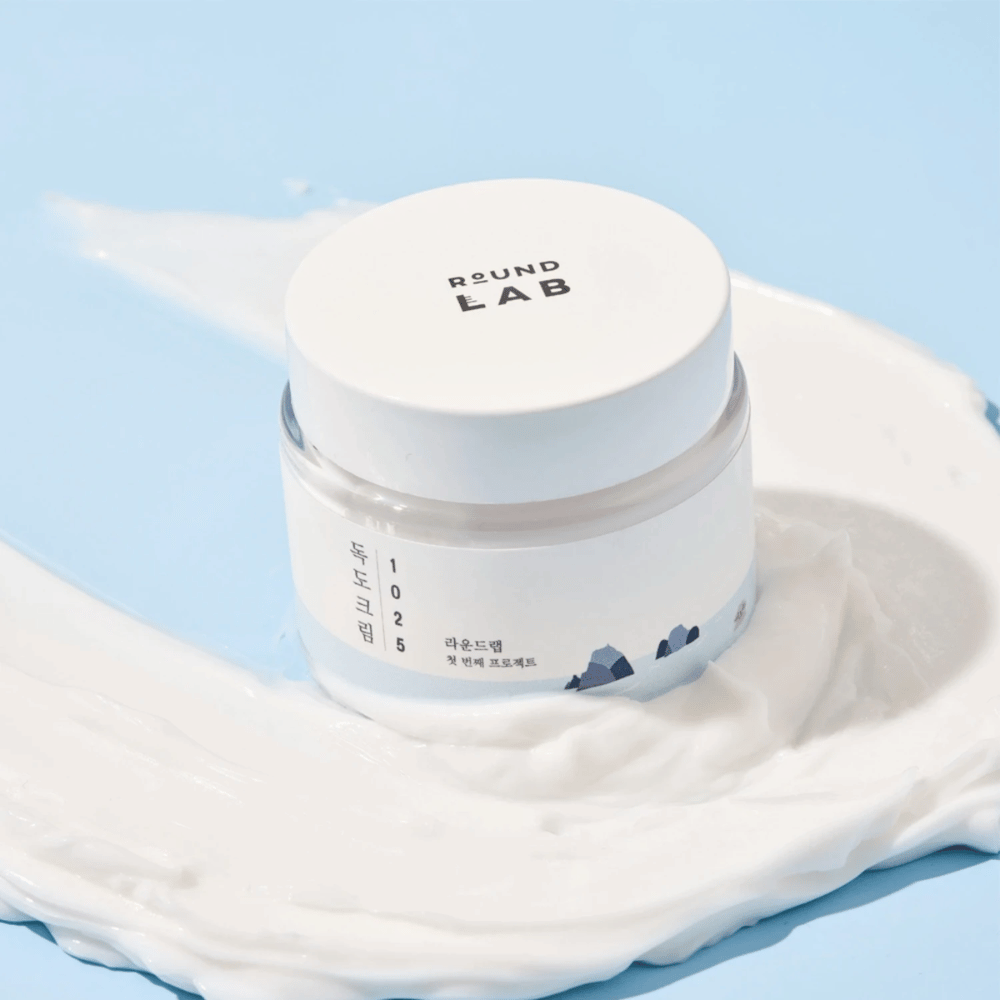 En krukke med Round Labs 1025 Dokdo Cream 80 ml som giver fugtgivning, placeret på en levende blå baggrund.