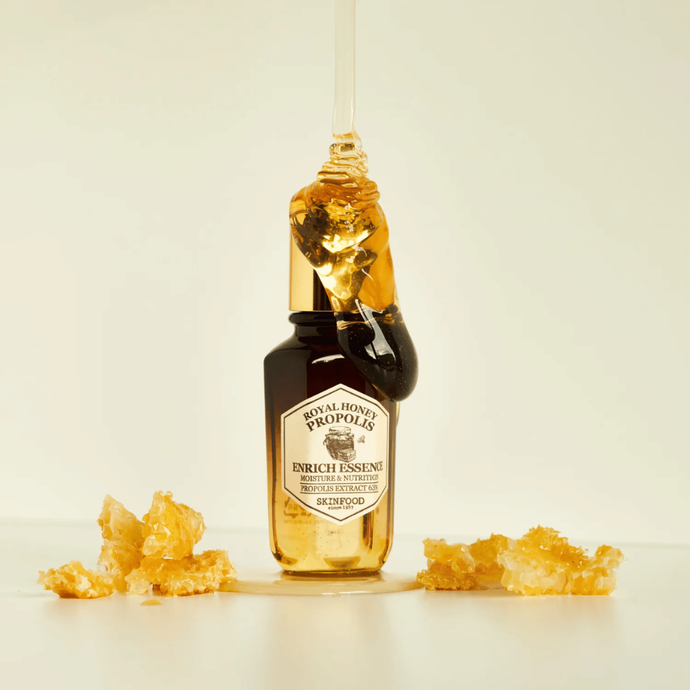 Billede af en glasflaske med Royal Honey Propolis Enrich Essence fra Skinfood. En stor dråbe af gylden honning løber ned fra flaskens guldfarvede låg. Rundt om flasken ligger små stykker af honningkage spredt på en lys overflade. Essensen markedsføres med fokus på fugt og næring, med 63% propolisekstrakt.