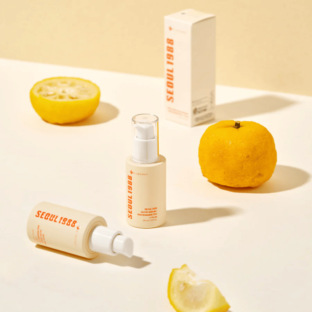 Flere flasker og emballager af Seoul 1988 Glow Serum arrangeret med hele og halverede yuja-frugter i baggrunden.