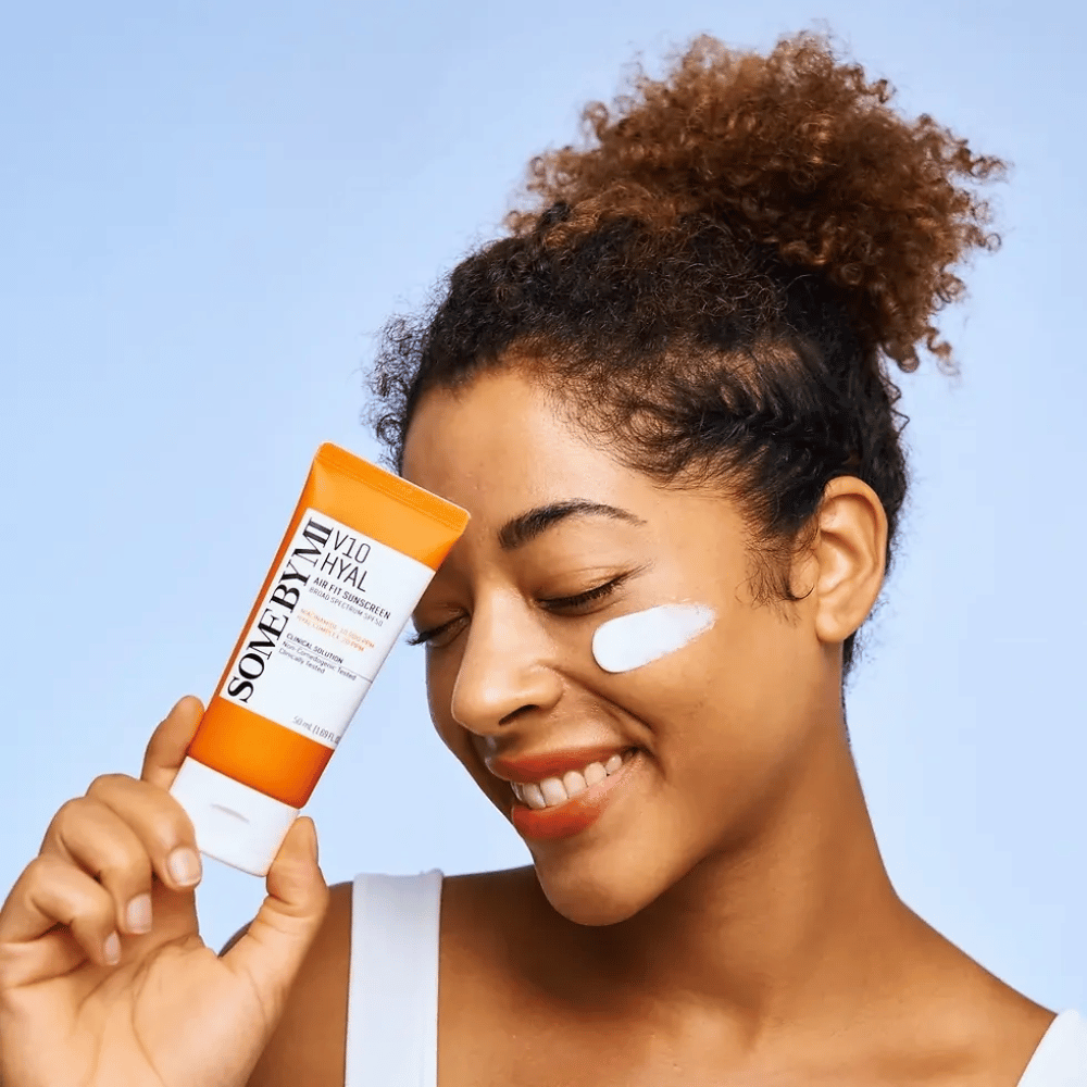 En kvinde holder en tube med SOME BY MI V10 Hyal Air Fit Sunscreen og har en hvid plet af solcreme på kinden. Hun smiler glad og fremviser tydeligt produktet, hvilket fremhæver dets anvendelse og fordele for hudpleje og beskyttelse mod solen.