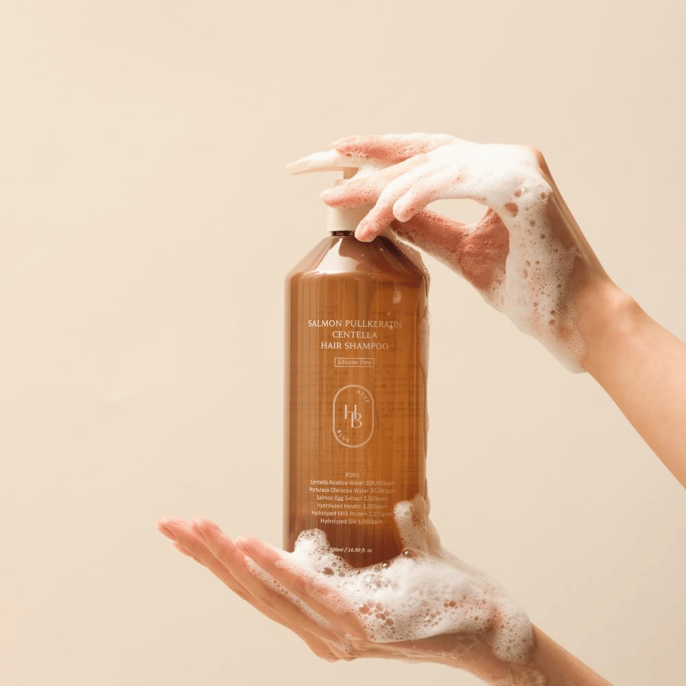 Heveblue Salmon Pullkeratin Centella Hair Shampoo i brun flaske omgivet af skum. Produktet holdes mellem to hænder og fremhæver en rensende og plejende hårplejeoplevelse med rigeligt skum.