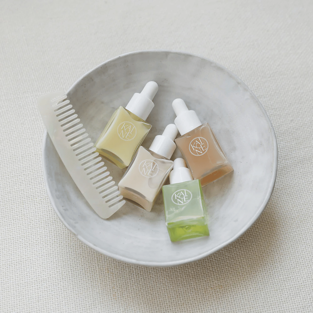 Fire små serumflasker fra KAINÉ Serum Discovery Kit ligger i en hvid skål ved siden af en kam, med en naturlig og rolig baggrund i beige tone.
