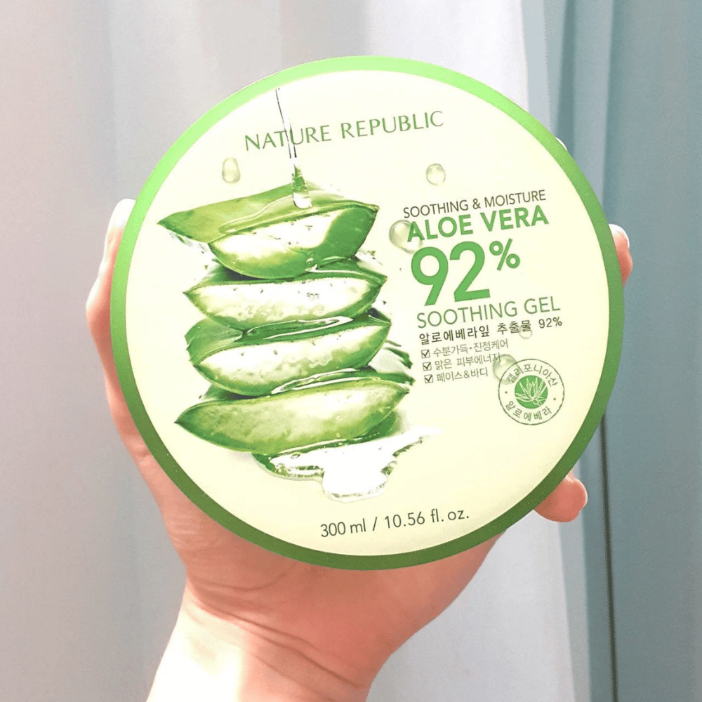 En hånd holder en rund bøtte med Nature Republic Aloe Vera 92% Soothing Gel. Bøtten er grøn med et billede af aloe vera og tekst, der beskriver dens beroligende og fugtgivende egenskaber. Indholdet er 300 ml.