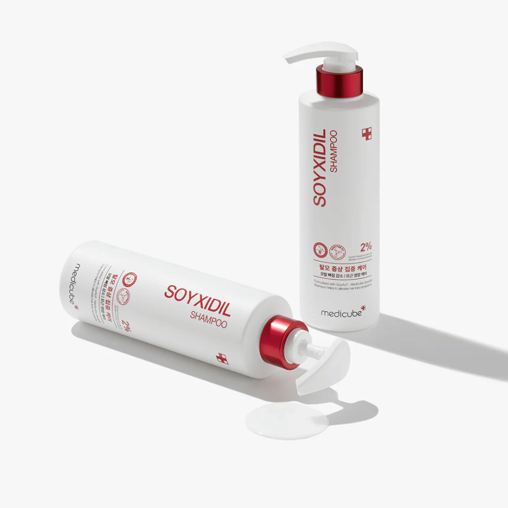 To flasker af Medicube Soyxidil Shampoo i hvid og rød design, en stående og en liggende med shampoo spildt ved siden af.